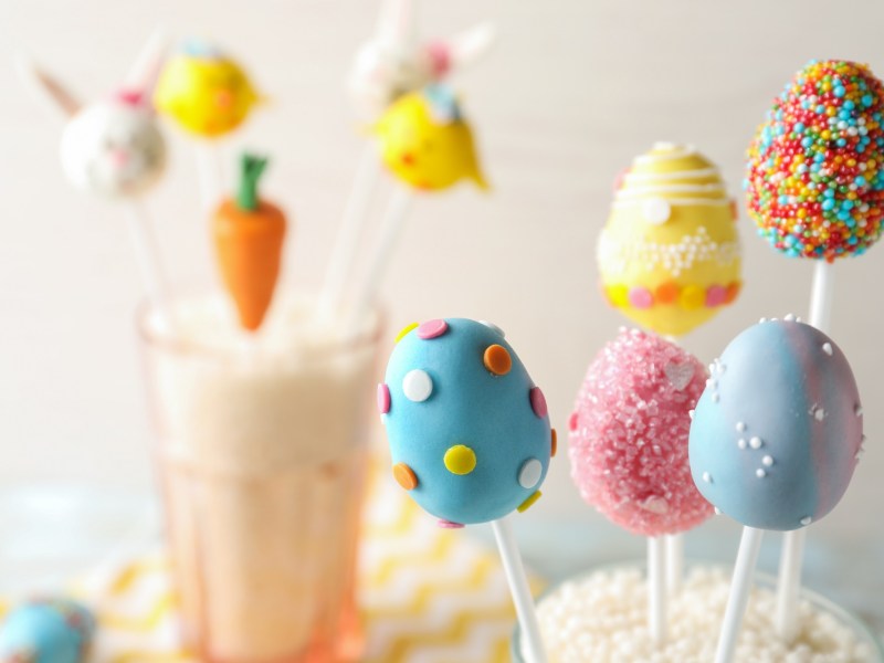 Bunte Oster-Cakepops am Stiel, verziert mit Zuckerperlen, im Hintergrund unscharf weitere Cakepops.