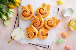 Mehrere Osterhasen-Zimtschnecken auf einem Kuchengitter, drumherum Tulpen und bunte Ostereier.