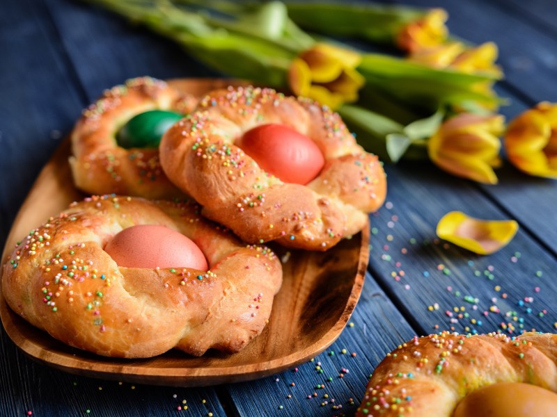 3 Osterkränze aus Hefeteig mit je einem bunten Ei in der Mitte auf einem Holzteller, daneben Osterglocken.