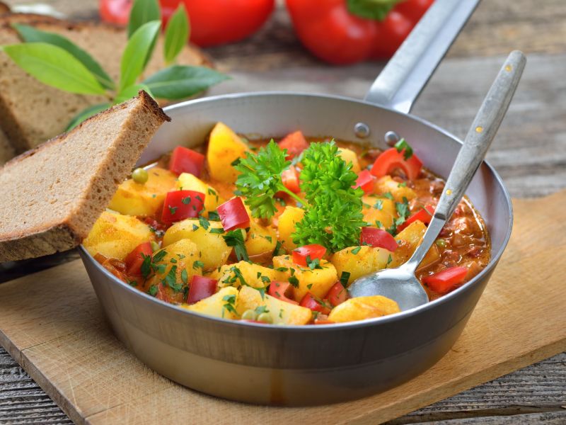 Paprika-Kartoffel-Topf, serviert in einer Pfanne. Ein Löffel steckt im Essen, eine Scheibe Brot liegt daneben.