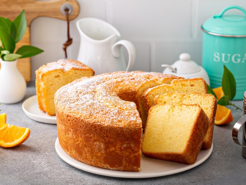 Ein angeschnittener Pfundkuchen auf einer hellgrauen Arbeitsfläche. Daneben stehen eine Zuckerdose, ein Krug und ein Puderzucker-Streuer.