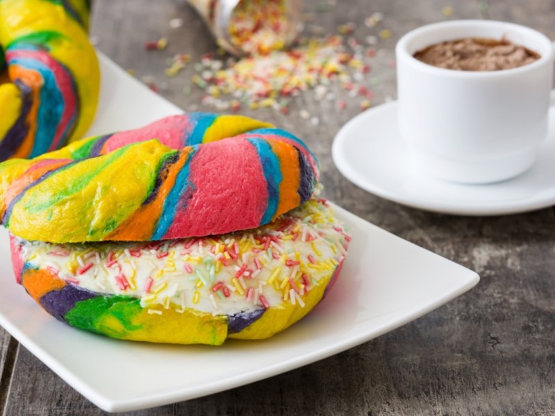 Ein aufgeschnittener Rainbow Bagel mit Frischkäsefüllung und Streuseln. Daneben steht eine Tasse Kakao.