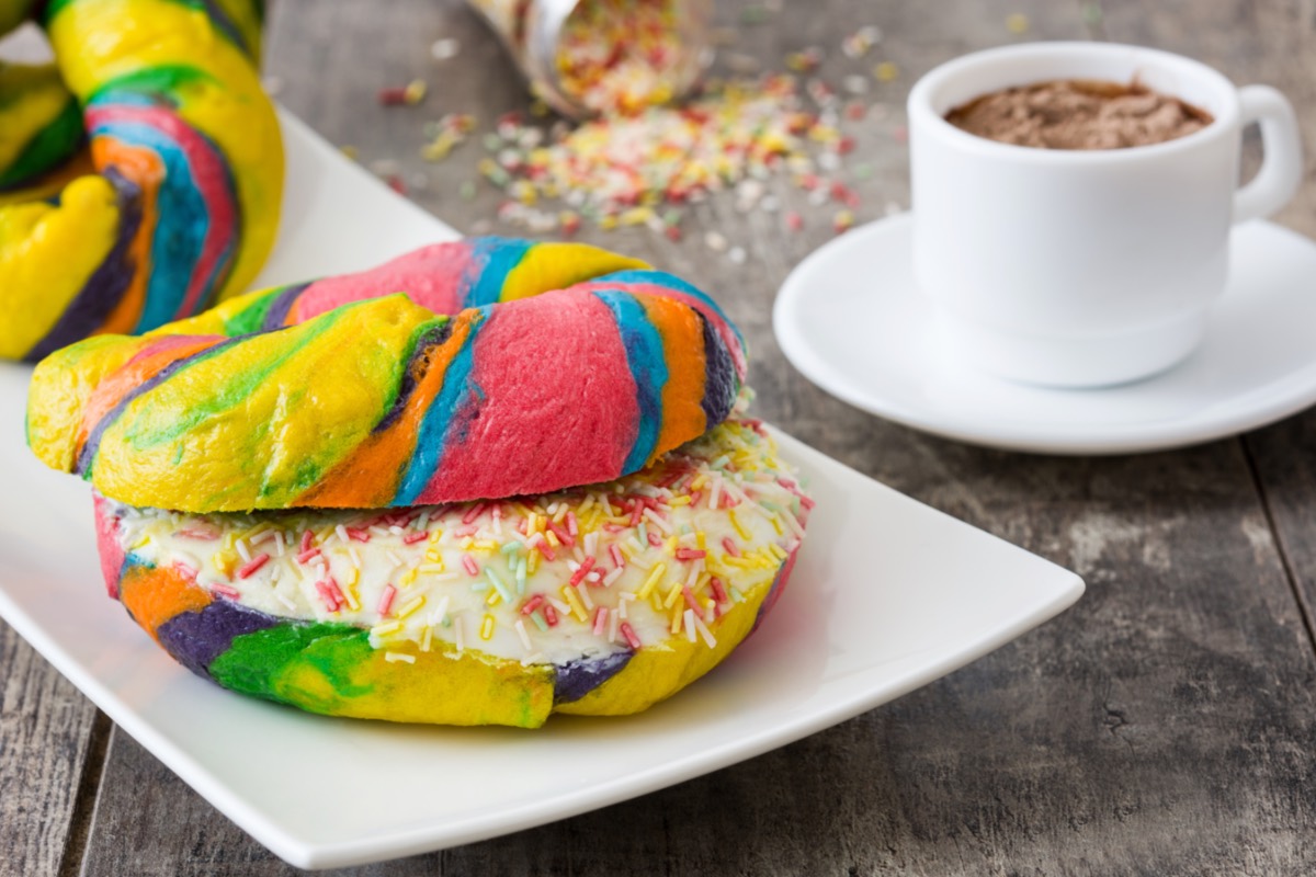 Ein aufgeschnittener Rainbow Bagel mit Frischkäsefüllung und Streuseln. Daneben steht eine Tasse Kakao.