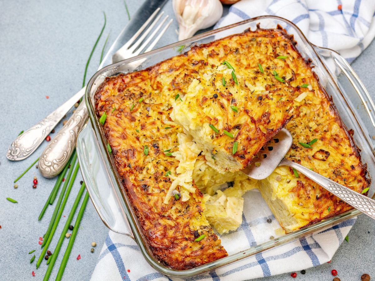 Rösti-Schnitzel-Auflauf in einer gläsernen Ofenform. Darunter liegt ein kariertes Küchentuch und Besteck sowie Frühlingszwiebeln als Deko daneben.