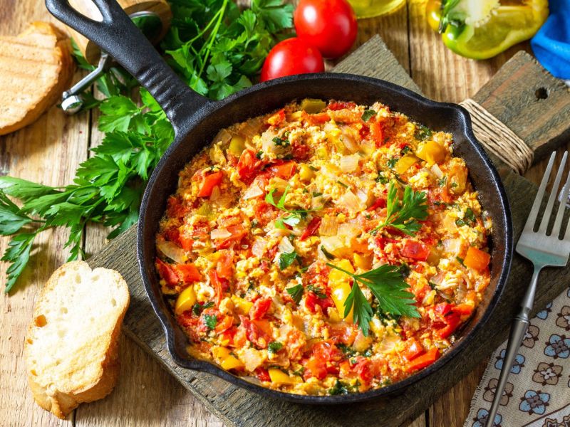 Rührei mit Gemüse, serviert in einer gusseisernen Pfanne. Im Hintergrund liegen Kräuter, kleine Tomaten und frisches Brot als Deko.