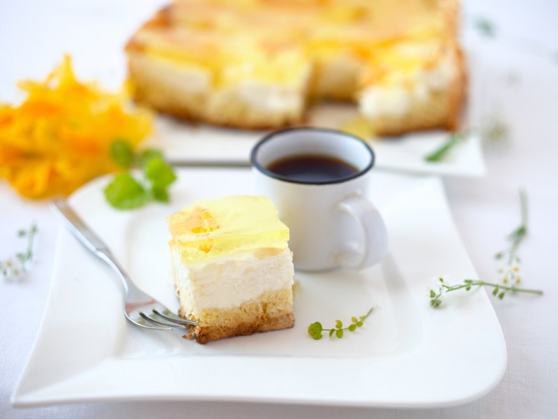 Ein Stück Schmankuchen vom Blech mit Früchten auf einem Teller, daneben eine Kaffeetasse, dahinter der Kuchen.