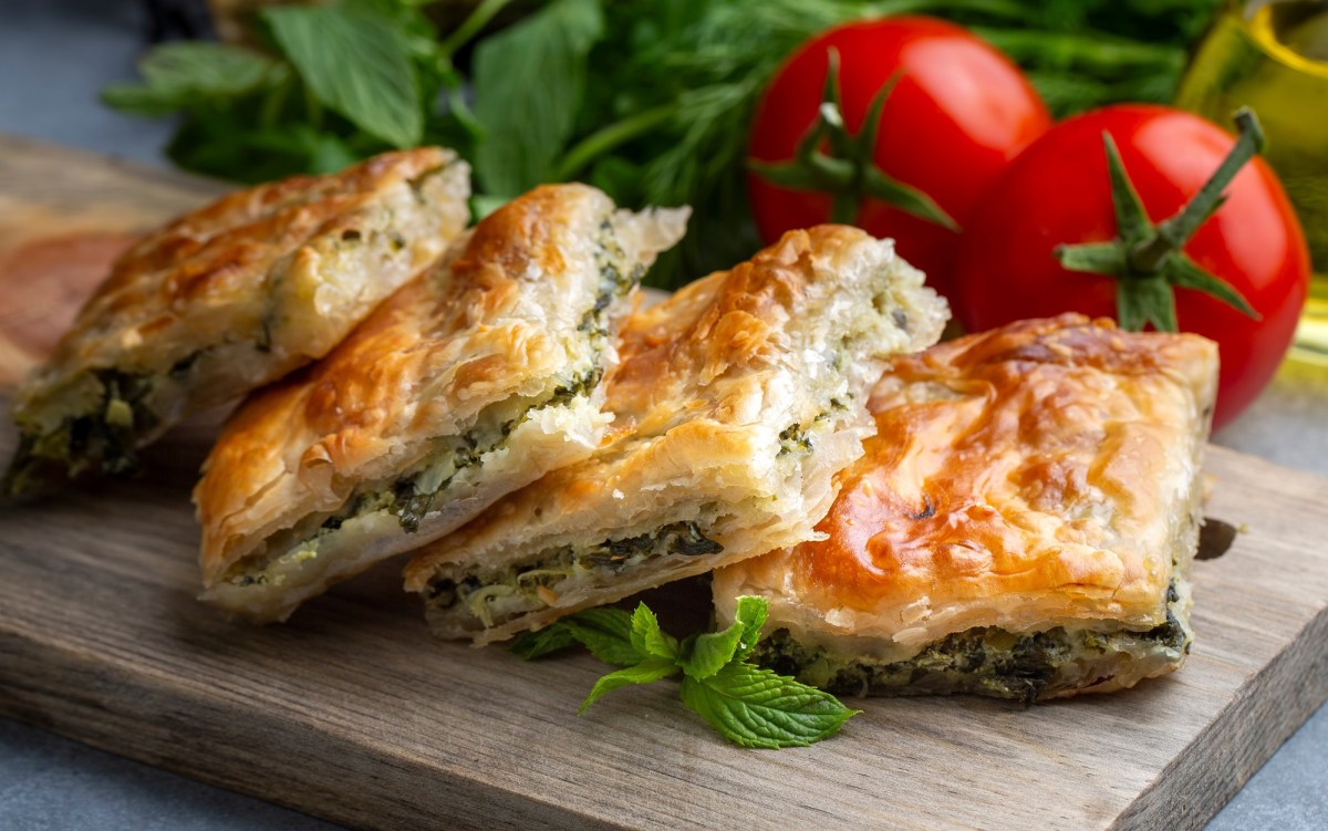 4 Stücke Spinat-Börek mit Feta auf einem Brett, dahinter Tomaten und frischer Spinat.