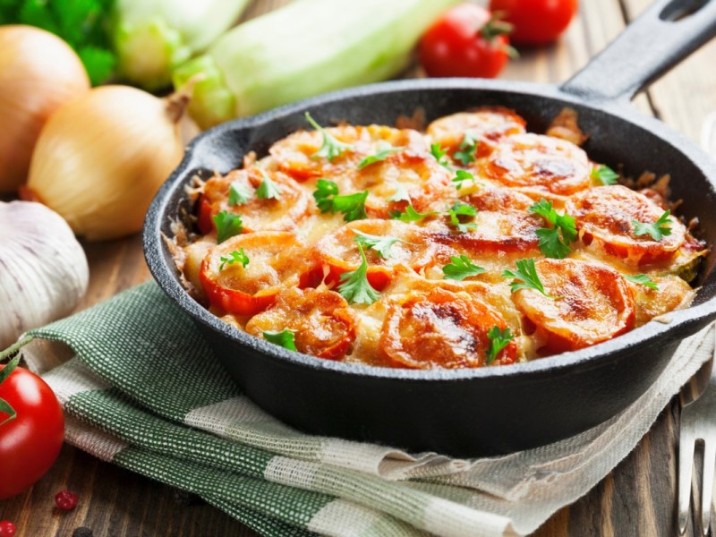 Tomaten-Zucchini-Auflauf in einer feuerfesten Pfanne auf einem Geschirrtuch. Im Hintergrund liegen frische Zutaten.