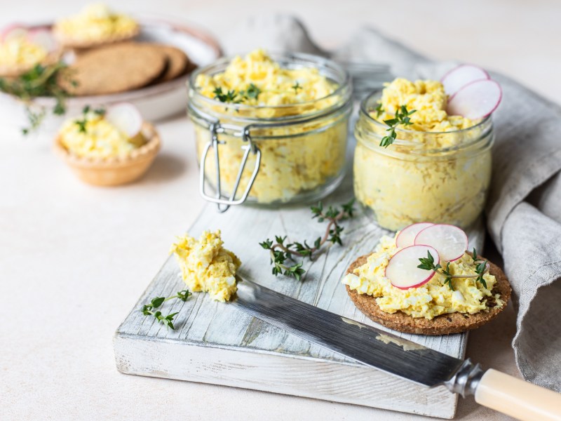 Zwei Gläser veganer Eiersalat auf einem Brett, darauf auch eine runde Scheibe Schwarzbrot mit Eiersalat und ein Messer.