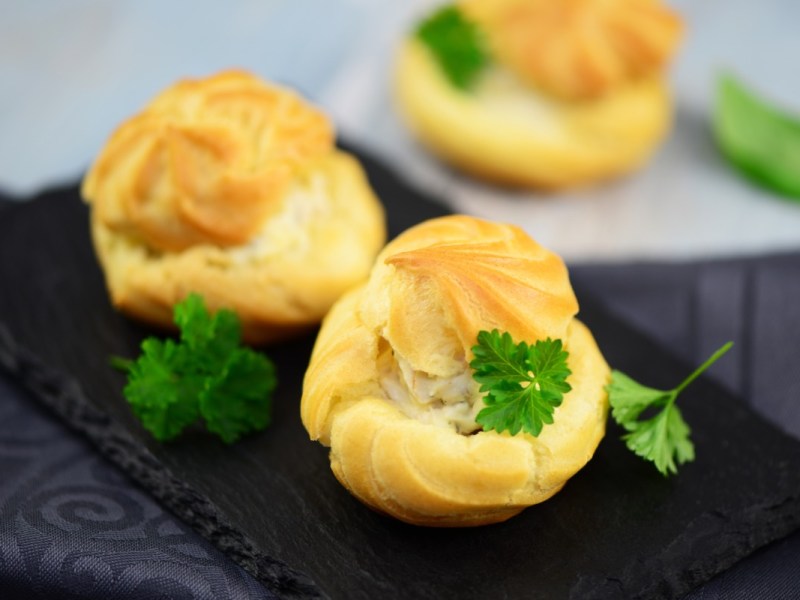 Drei Windbeutel mit Frischkäse-Füllung auf einer Schiefertafel. Garniert sind sie mit Petersilie.