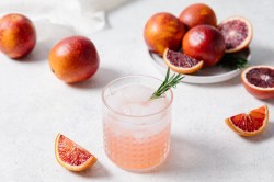 Ein Glas Aperol Paloma mit Eiswürfeln und Grapefruits daneben.