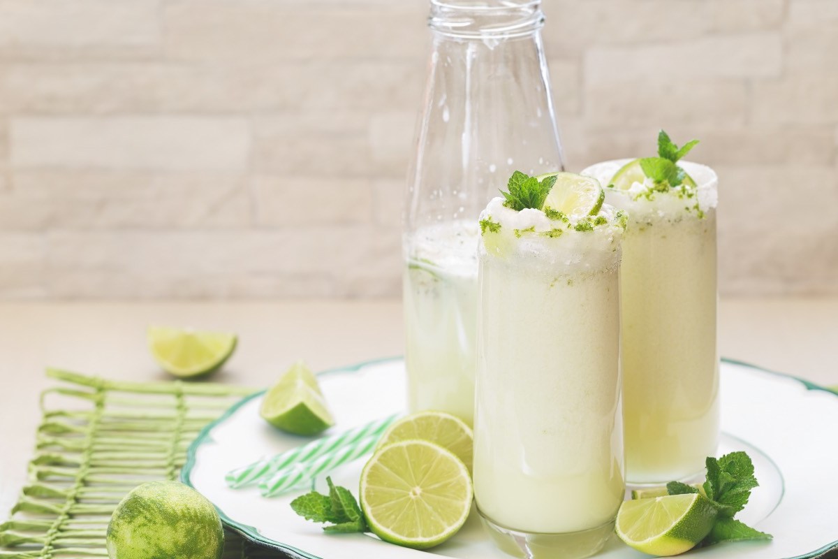 2 Gläser brasilianische Limonade neben einer Flasche auf einem runden Tablett. Daneben liegen Limettenscheiben und Minzblätter.