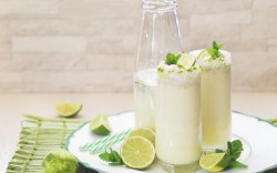 2 Gläser brasilianische Limonade neben einer Flasche auf einem runden Tablett. Daneben liegen Limettenscheiben und Minzblätter.