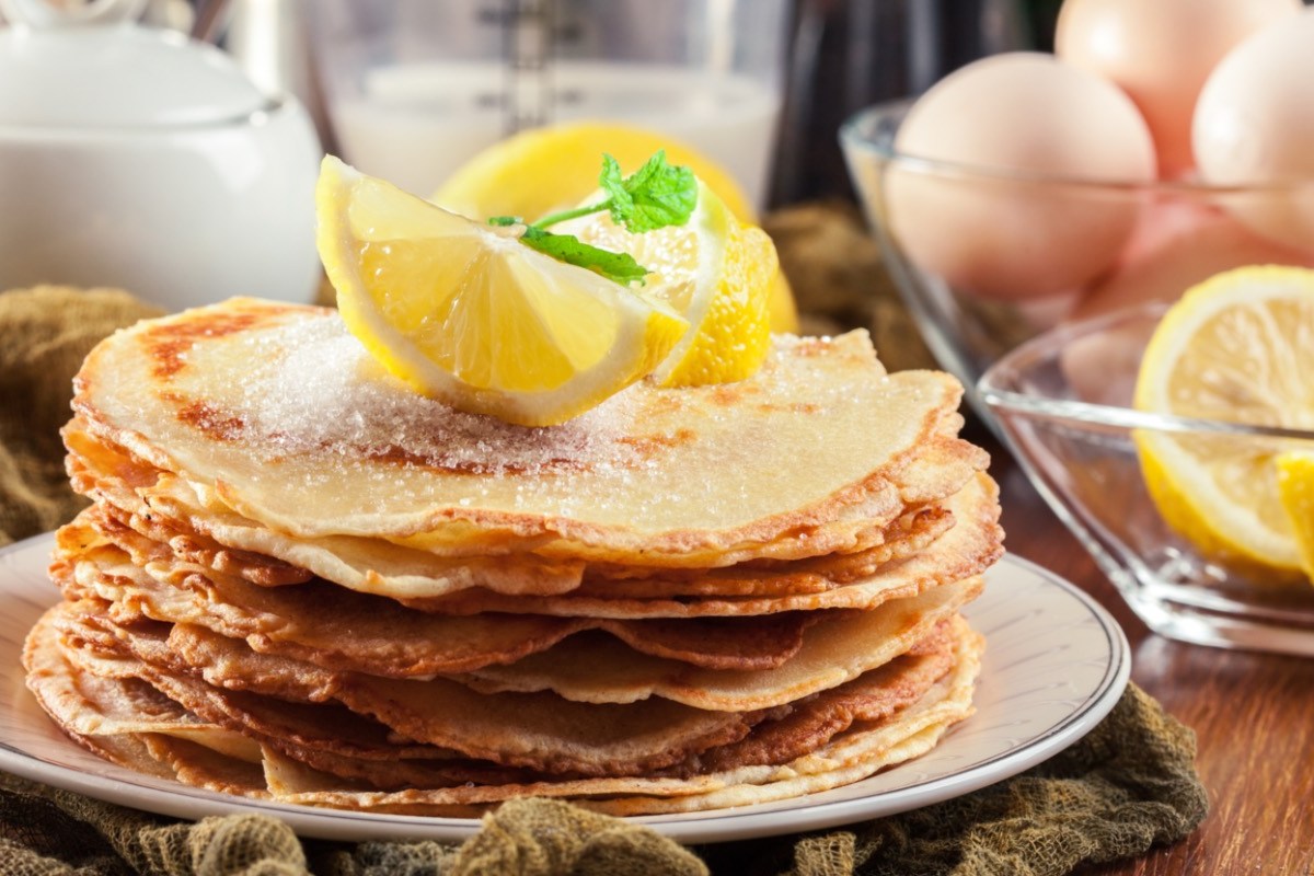 Ein Stapel britische Pancakes mit Zitronenstücken obendrauf. Im Hintergrund stehen die Zutaten.