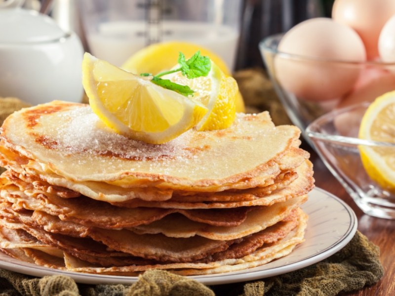 Ein Stapel britische Pancakes mit Zitronenstücken obendrauf. Im Hintergrund stehen die Zutaten.
