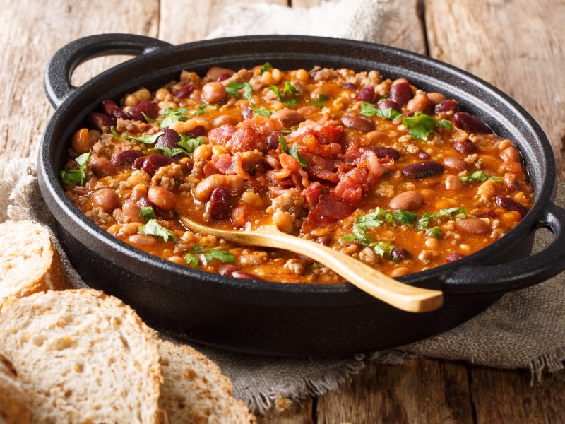 Cowboypfanne mit Kartoffeln in einem gusseisernen Topf. Darin steckt ein Holzlöffel und serviert ist es mit Brot.