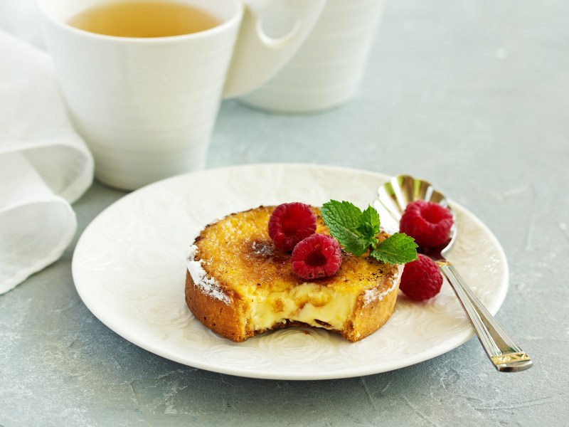 Crème-Brûlée-Kuchen, garniert mit Himbeeren und Minze, auf einem weißen Teller. Im Hintergrund steht eine Tasse Tee.