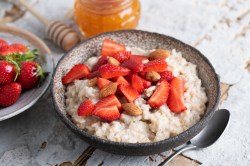 Eine dunkle Schüssel Erdbeer-Porridge, garniert mit Erdbeeren und ganzen Mandeln, daneben ein Teller mit Erdbeeren und ein Löffel.