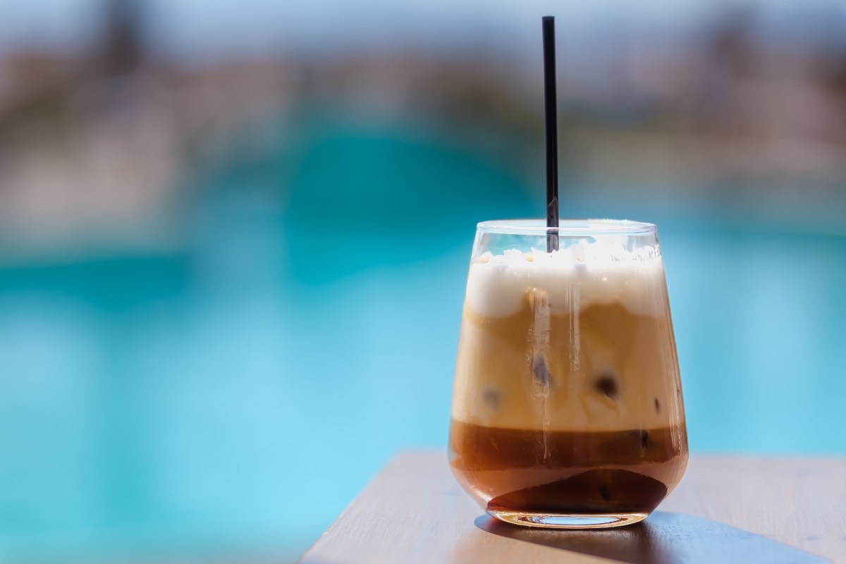 Ein Freddo Cappuccino in einem Glas mit Strohhalm. Im Hintergrund schimmert das Blau eines Pools.