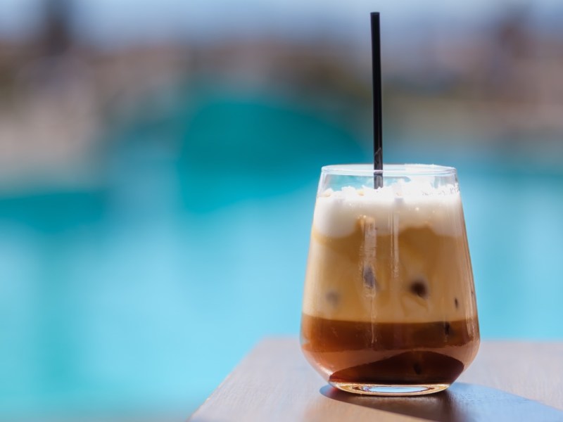 Ein Freddo Cappuccino in einem Glas mit Strohhalm. Im Hintergrund schimmert das Blau eines Pools.