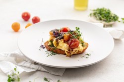 Ein Stück Frühstücks-Bruschetta mit Rührei, gerösteten Tomaten und Kresse auf einem Teller, daneben ein paar Tomaten und Kräuter.