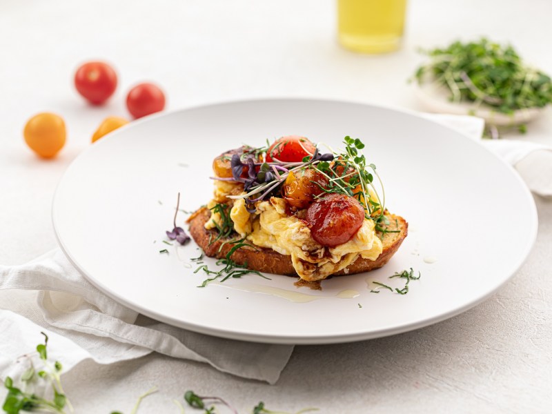 Ein Stück Frühstücks-Bruschetta mit Rührei, gerösteten Tomaten und Kresse auf einem Teller, daneben ein paar Tomaten und Kräuter.