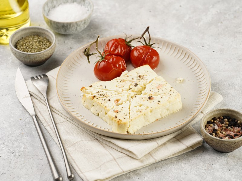 Gebackener Schafskäse auf einem Teller, daneben gebackene Tomaten mit Strunk, Besteck, eine Schale Salz und Pfeffer sowie eine Flasche Öl.