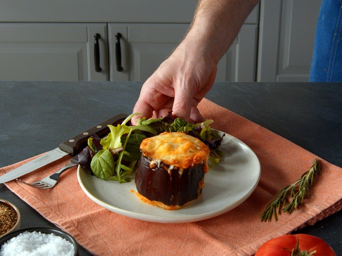 Gefüllte Auberginen-Becher: Eine Portion auf einem Teller, angerichtet mit Salat. Eine Hand präsentiert das Gericht.