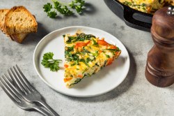 Ein Stück Gemüse-Tortilla auf einem weißen Teller, garniert mit Petersilie. Daneben liegen einige Scheiben Brot und es steht eine Pfeffermühle. Im Hintergrund ist der Rest Tortilla zu erahnen.