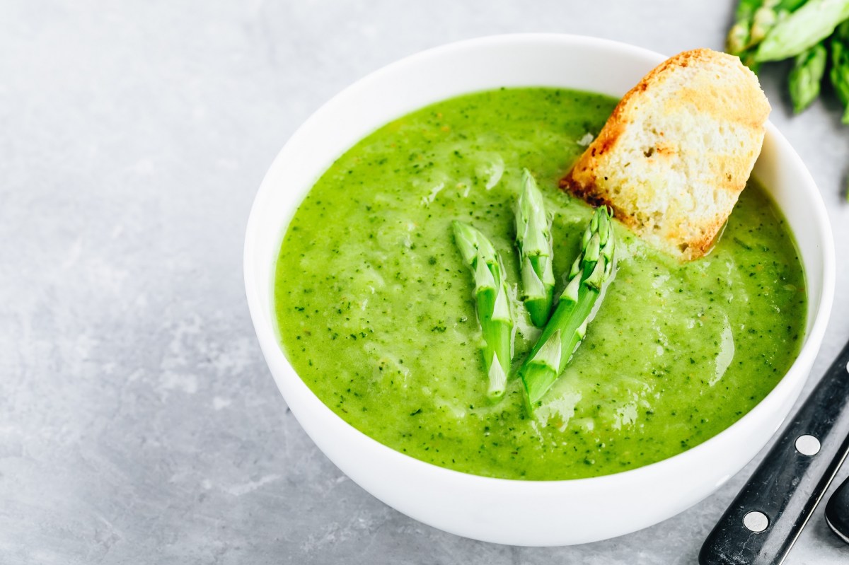 Grüne Spargelsuppe in einer weißen Schale, serviert mit Brot. Auf der Suppe liegen zwei Köpfe von grünem Spargel.