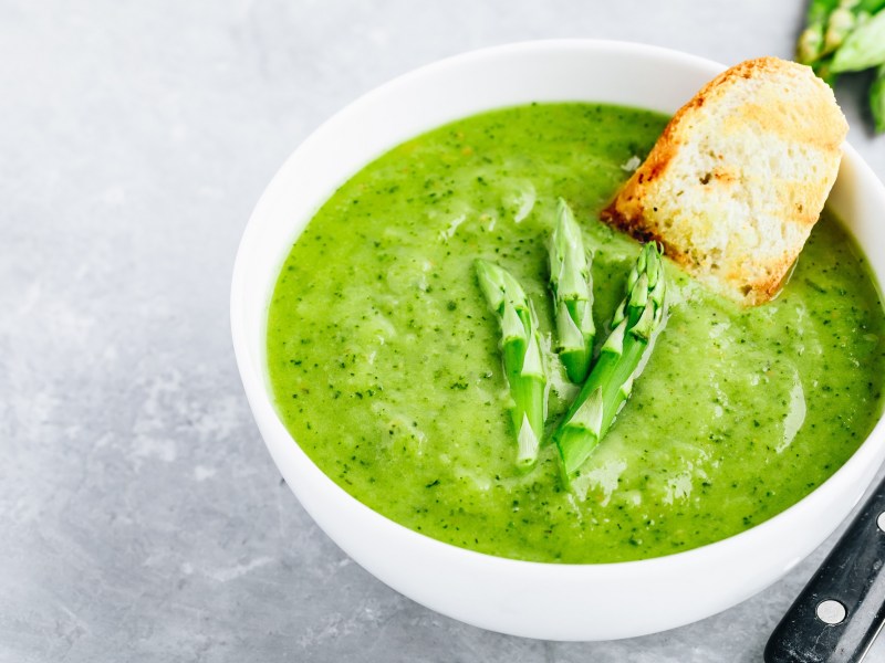 Grüne Spargelsuppe in einer weißen Schale, serviert mit Brot. Auf der Suppe liegen zwei Köpfe von grünem Spargel.