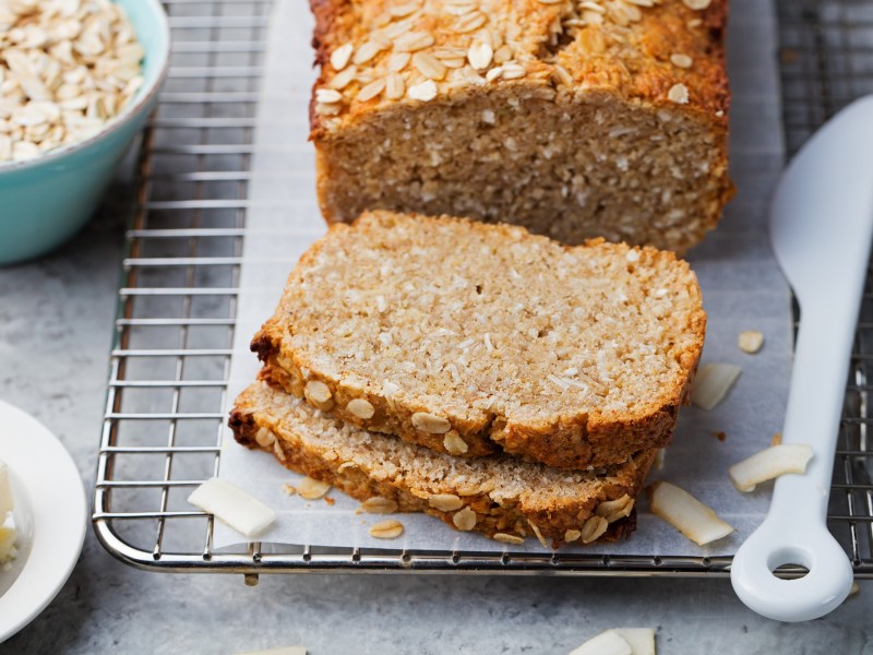 angeschnittenes Haferflocken-Quark-Brot auf einem Brett, daneben eine Schüssel Haferflocken und ein Messer.