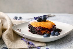 Eine Portion Heidelbeer-Auflauf mit frischen Blaubeeren auf einem Teller, daneben drapiert eine Stoffserviette und Blüten.