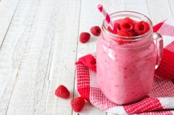 Ein Maison Jar Glas mit Himbeer-Joghurtdrink, garniert mit Himbeeren, darin ein Strohhalm, drumherum frische Beeren und ein kariertes Geschirrtuch.