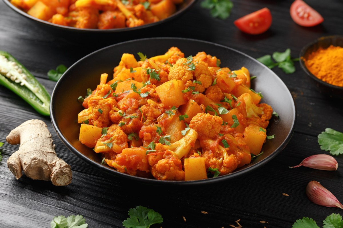 Ein Teller Indische Kartoffel-Blumenkohl-Pfanne, drumherum Zutaten wie Ingwer und Currypulver.
