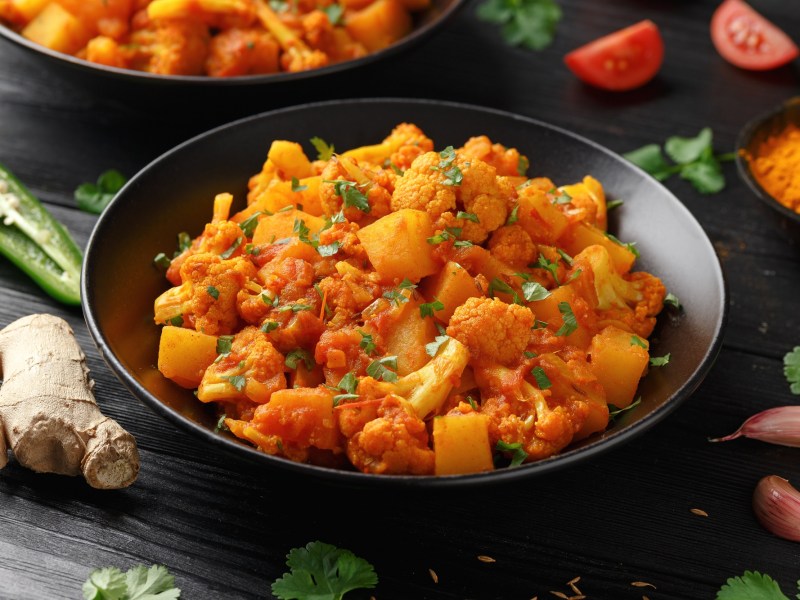 Ein Teller Indische Kartoffel-Blumenkohl-Pfanne, drumherum Zutaten wie Ingwer und Currypulver.