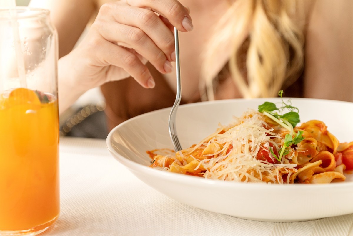 Jeden Tag Nudeln essen: eine blonde Frau wickelt mit einer Gabel Spaghetti in Tomatensoße mit Parmesan auf einem weißen Teller auf, daneben ein Glas Multivitamin-Saft, man sieht nur ihren Oberkörper, nicht ihr Gesicht.