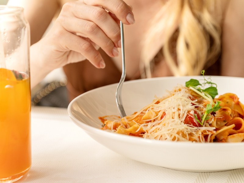Jeden Tag Nudeln essen: eine blonde Frau wickelt mit einer Gabel Spaghetti in Tomatensoße mit Parmesan auf einem weißen Teller auf, daneben ein Glas Multivitamin-Saft, man sieht nur ihren Oberkörper, nicht ihr Gesicht.