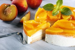 Käse-Sahne-Torte mit Pfirsichen, aus der ein Stück mit einem Tortenheber herausgenommen wird, daneben frische Pfirsiche.