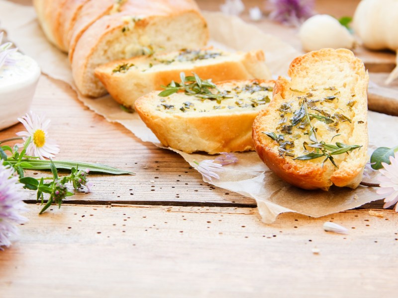 ein angeschnittenes selbst gemachtes Kräuterbaguette, daneben frische Wiesenblumen