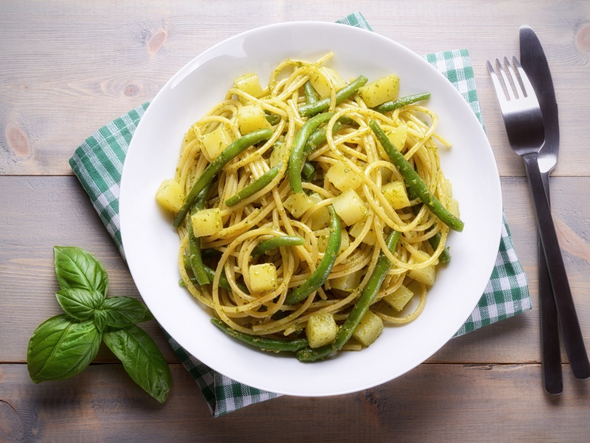 Ligurische Pasta mit Bohnen und Kartoffeln auf einem weißen Teller. Daneben liegt Besteck und etwas Basilikum.