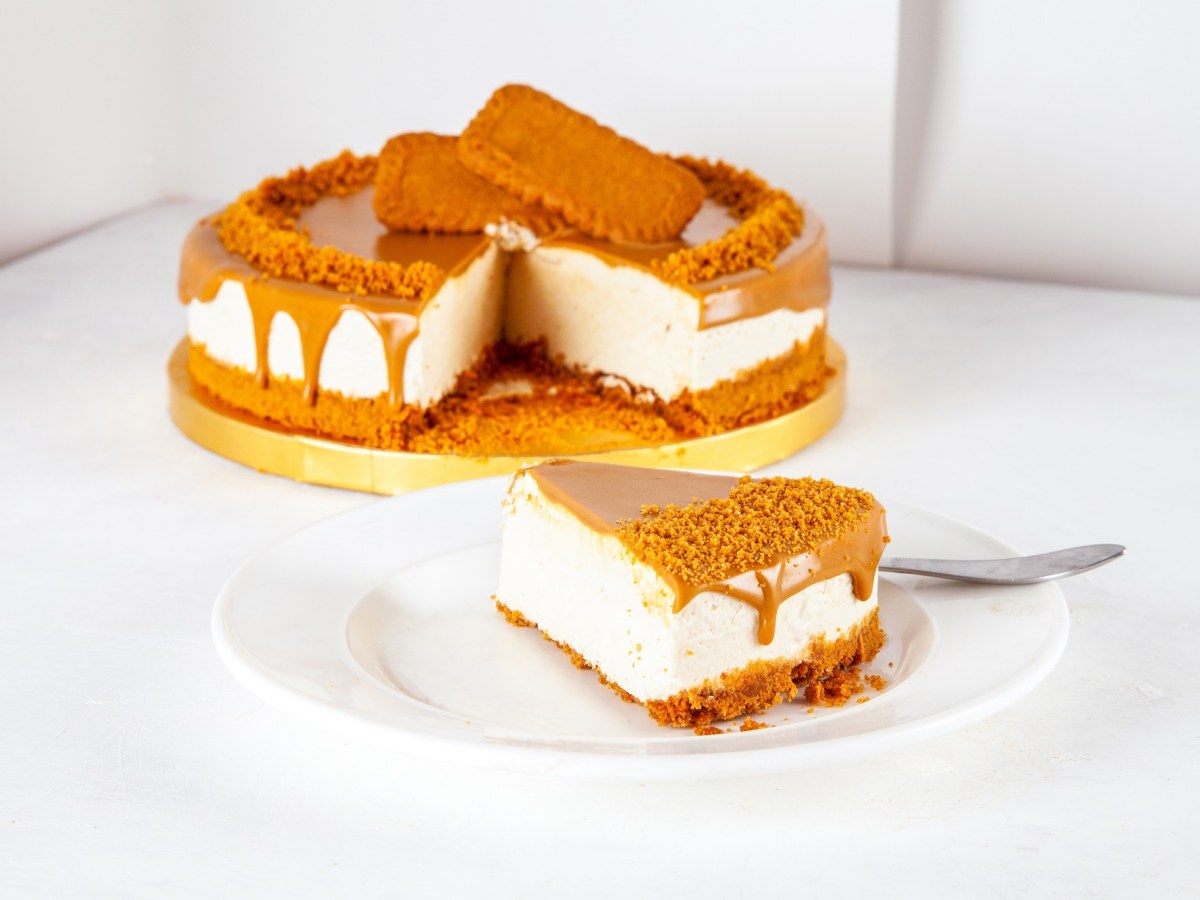 Ein Stück Lotus Cheesecake auf einem weißen Teller. Im Hintergrund steht der Rest des Kuchens auf einer gelben Servierplatte
