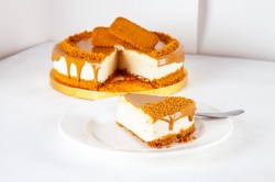 Ein Stück Lotus Cheesecake auf einem weißen Teller. Im Hintergrund steht der Rest des Kuchens auf einer gelben Servierplatte