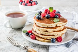 Ein Stapel Quark-Pancakes auf einem Teller, dekoriert mit frischen Beeren, daneben Besteck und eine Kaffeetasse.