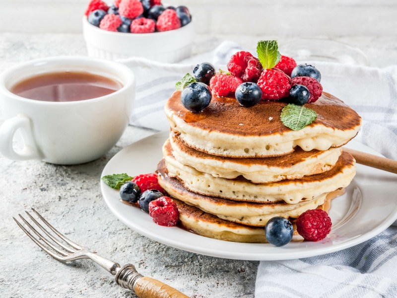 Ein Stapel Quark-Pancakes auf einem Teller, dekoriert mit frischen Beeren, daneben Besteck und eine Kaffeetasse.