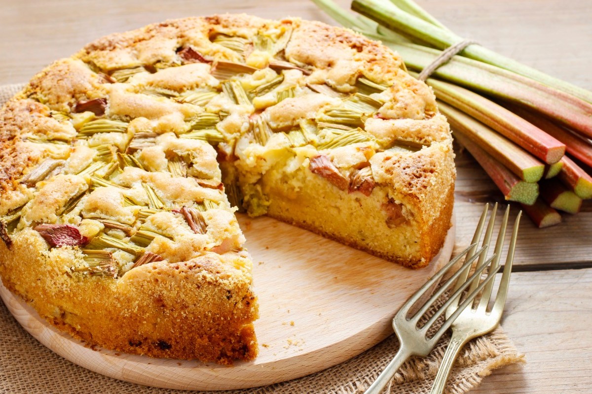 Ein Rhabarber-Apfel-Kuchen auf einem runden Holzbrett, aus dem ein Stück herausgeschnitten wurde. Daneben liegen Besteck und Rhabarberstangen.