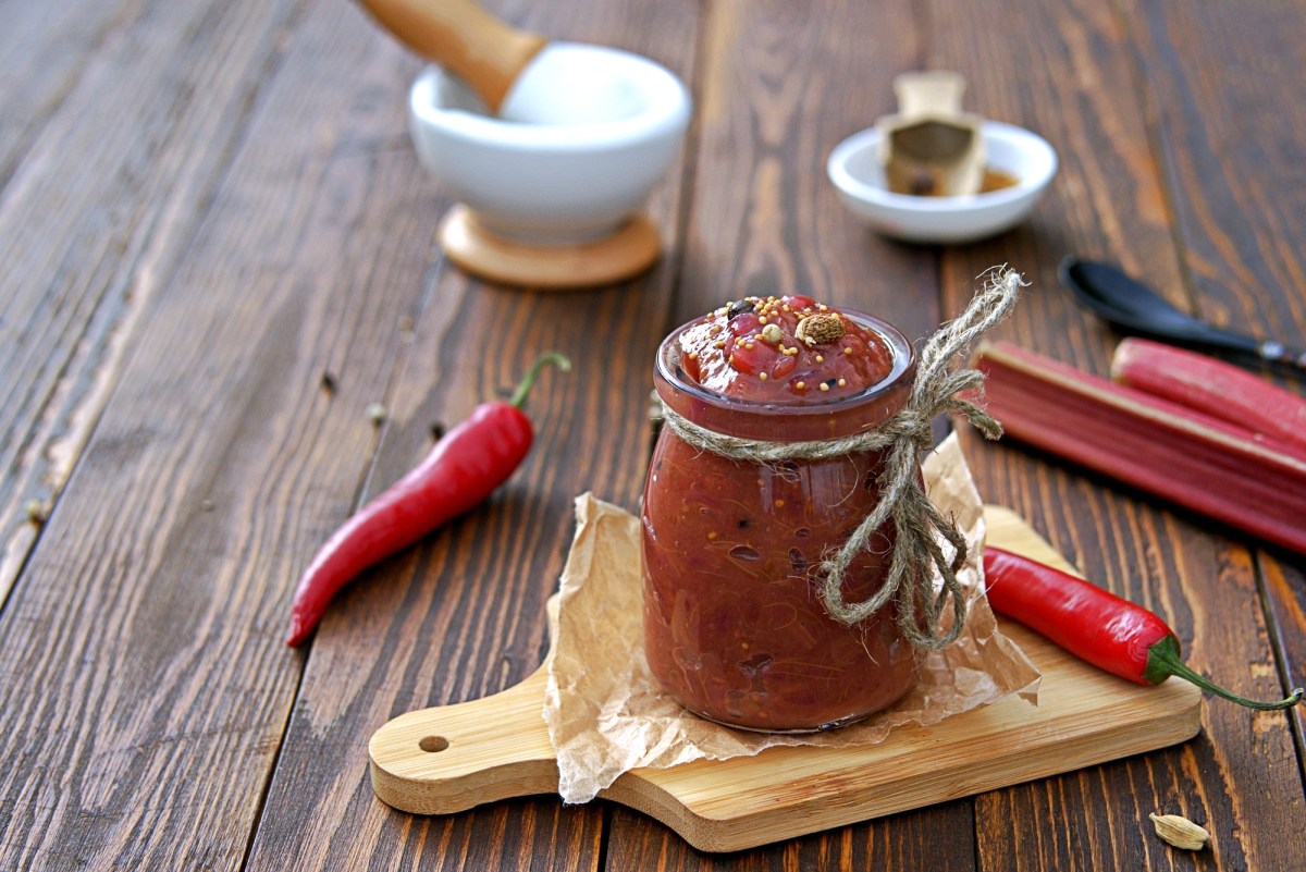 Glas mit Rhabarber-Chutney auf Brett, Chilis und Rhabarber daneben.