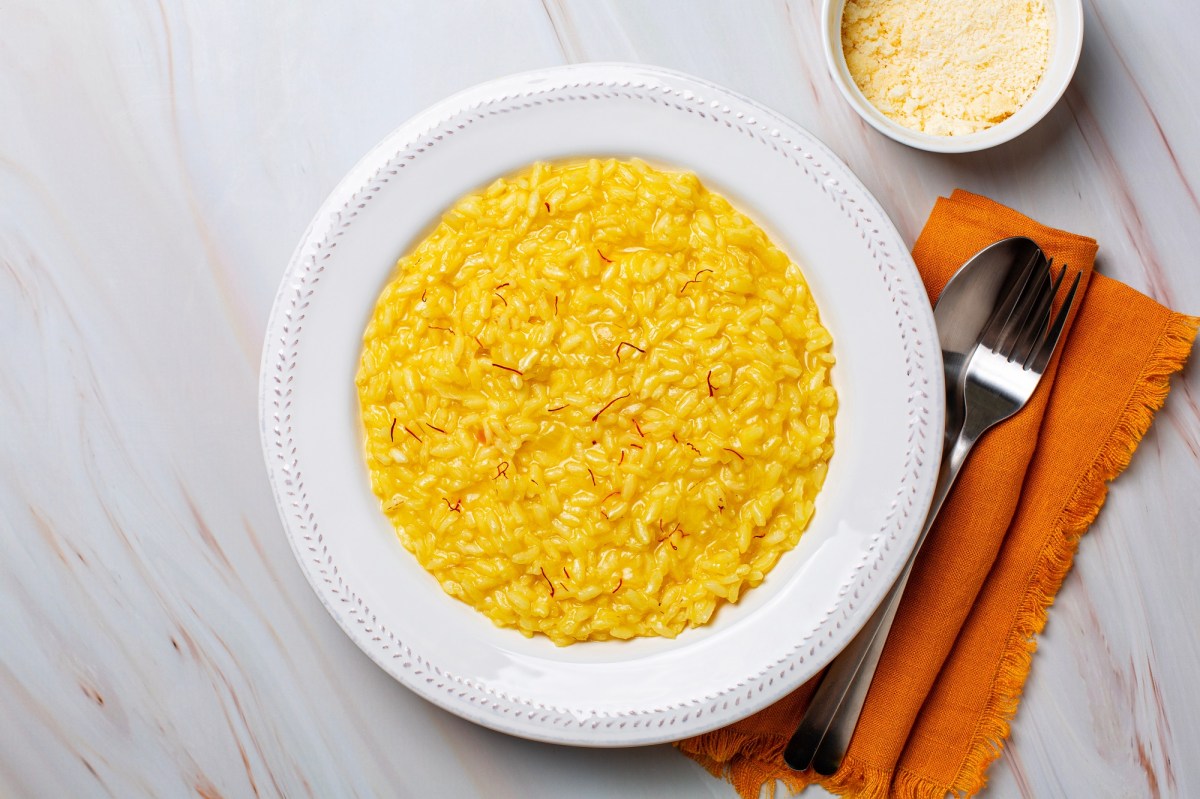 Risotto alla Milanese in einem weißen Teller. Daneben liegen Gabel und Löffel sowie eine orange Serviette. Eine Schale mit Parmesankäse steht auch dabei.