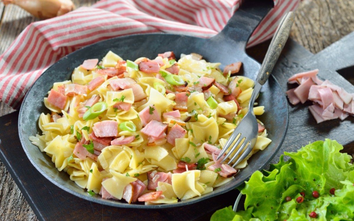 Schinkenfleckerl in einer Pfanne. Daneben ein Salat.