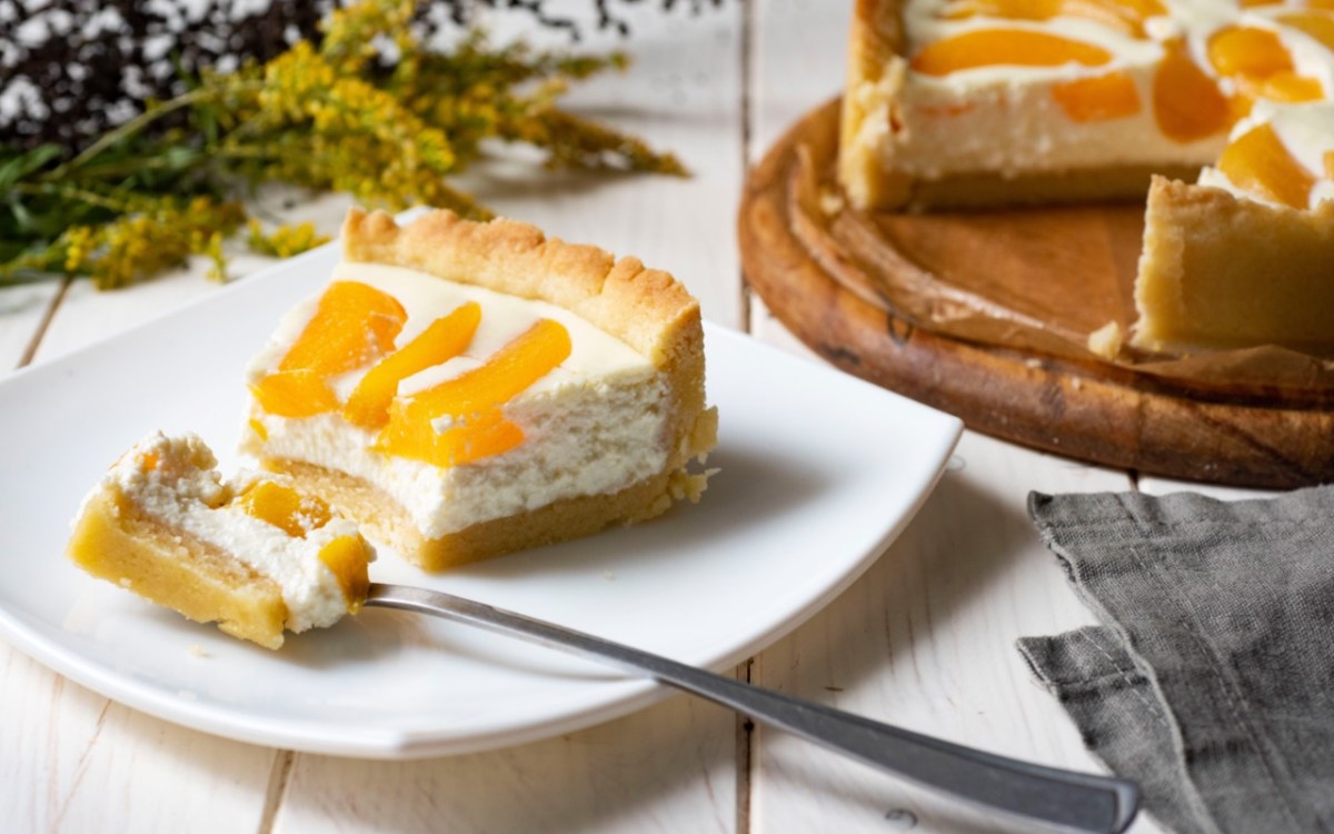 Ein Stück Schmandkuchen mit Pfirsichen auf einem Teller. Im Hintergrund steht der restliche Kuchen.